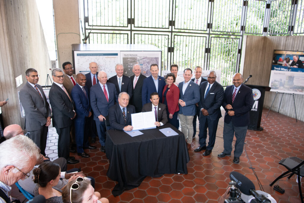 County executives gathered together. Two males seated in front, holding up signed paperwork.
