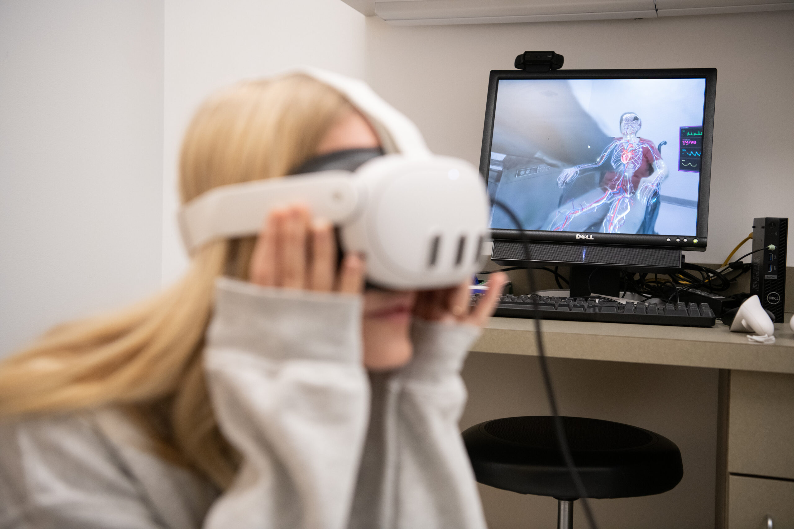 Blurry female with virtual reality goggles on stands with clear computer and human pictured in chair on screen in the background.