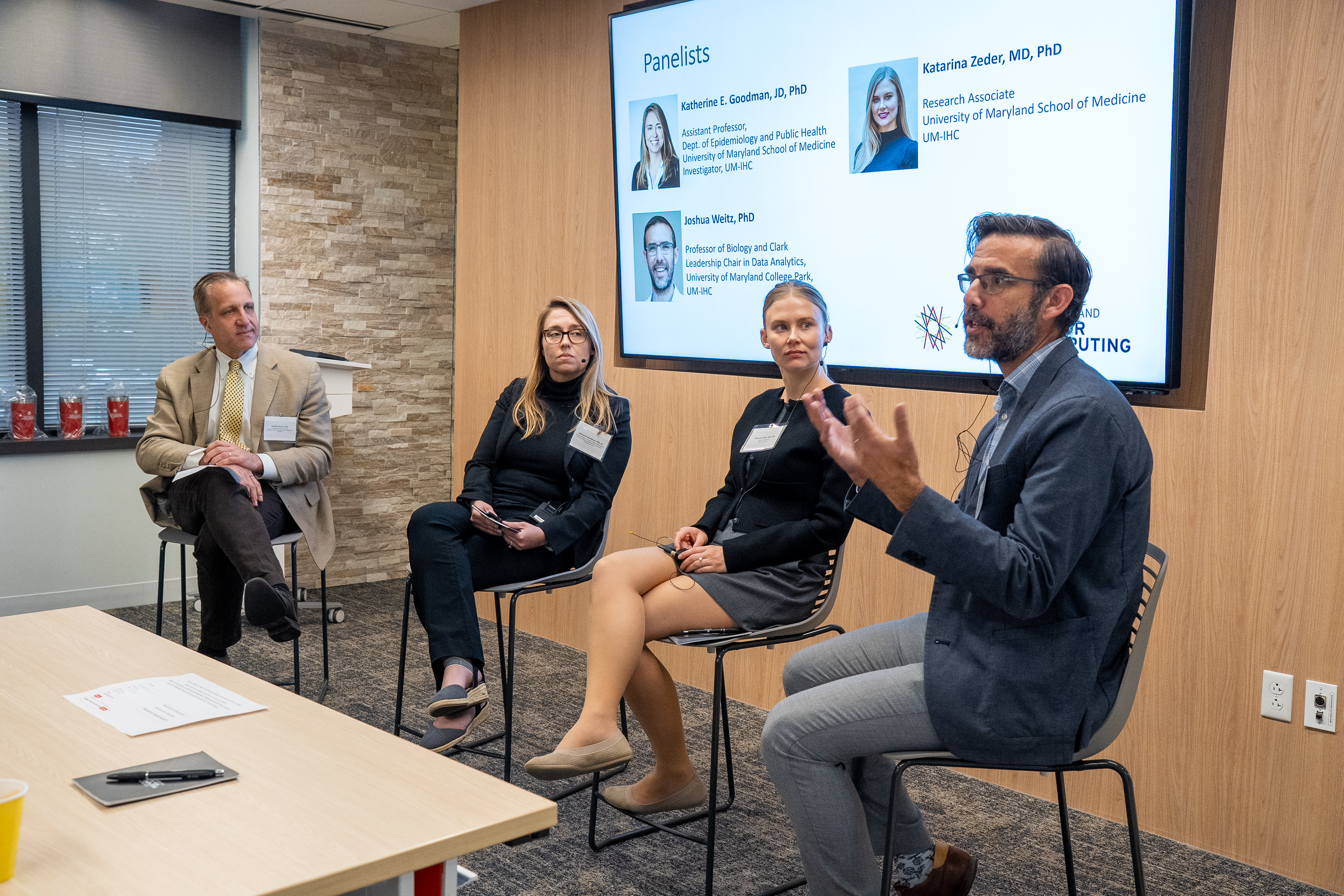 Four panelists seated in front of a screen, in the front of the room. Male panelist all the way to the left speaks.