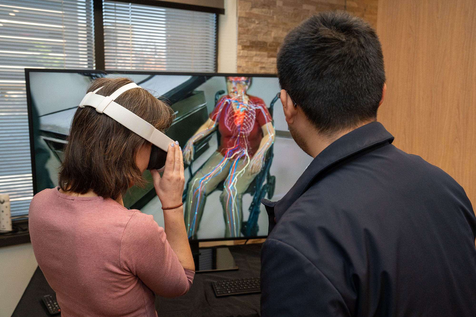 Male and female with the back of their heads to us, standing in front of TV monitor. Female is wearing virtual reality goggles.